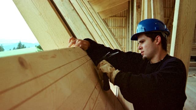 Ein Schreinerlehrling hämmert an einem Holzbrett, er trägt einen Handschuh, einen blauen Helm und einen schwarzen Pullover. Ein Schreinerlehrling hämmert an einem Holzbrett, er trägt einen Handschuh, einen blauen Helm und einen schwarzen Pullover.
