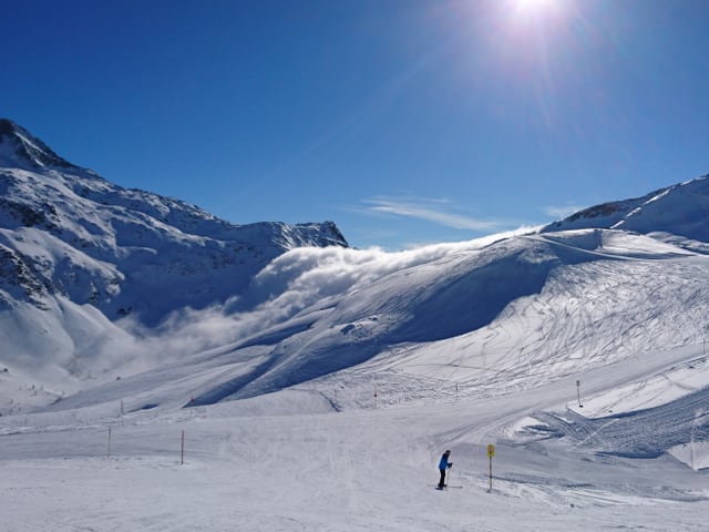 Verschneite Pisten, dahinter Nebelfall, darüber blauer Himmel.