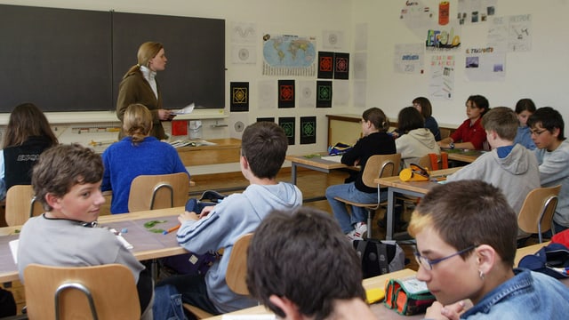 Blick in ein Schulzimmer: Die Lehrerin steht vor der Wandtafel und spricht mit den Schülerinnen und Schülern.