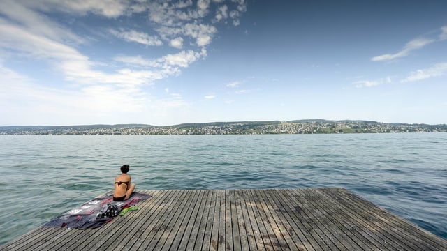 Eine Frau sitzt auf einem alten Holzfloss und blickt auf den blauen Zürichsee. Der Himmel ist mit leichten Wolken behangen. Eine Frau sitzt auf einem alten Holzfloss und blickt auf den blauen Zürichsee. Der Himmel ist mit leichten Wolken behangen.