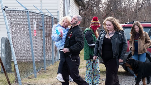 Szene auf einem Parkplatz vor einem Haus, im Hintergrund ein Auto: Ein Mann mit Tattoos im Gesicht trägt ein kleines Mädchen auf dem Arm. Hinter den beiden gehen eine Frau und zwei ältere Mädchen.