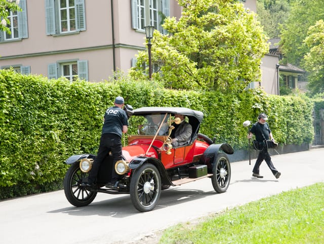 Kamermann steht auf Autohaube, hinten Tontechniker, Auto fährt an Villa vorbei