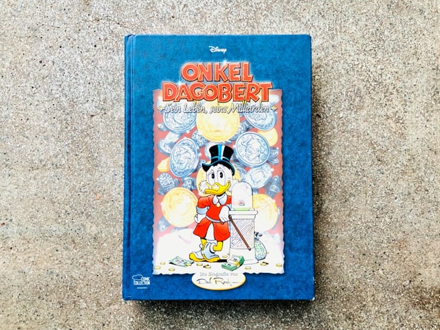 Der Comic-Sammelband «Onkel Dagobert – Sein Leben, seine Milliarden» geschrieben und gezeichnet von Don Rosa liegt auf einem Zementboden Der Comic-Sammelband «Onkel Dagobert – Sein Leben, seine Milliarden» geschrieben und gezeichnet von Don Rosa liegt auf einem Zementboden