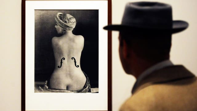 Mann mit Hut steht vor dem Foto eines Frauenrückens auf den die Schalllöcher einer Geige gemalt sind. Mann mit Hut steht vor dem Foto eines Frauenrückens auf den die Schalllöcher einer Geige gemalt sind.