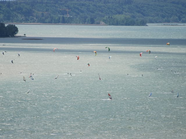 Viel Seeoberfläche von oben gesehen, auf dem Wasser farbige Punke, es sind Kiter und Windsurfer.
