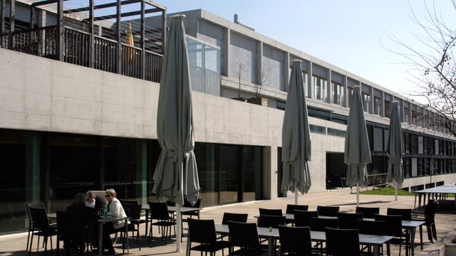 ein Betongebäude mit grossen Fenstern, davor eine Terrasse mit Tischen und Stühlen ein Betongebäude mit grossen Fenstern, davor eine Terrasse mit Tischen und Stühlen
