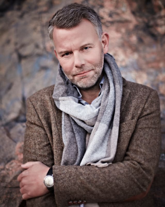 Ein Mann mit graumellierten Haaren und um den Hals geschwungenen Schal blickt in die Kamera. Ein Mann mit graumellierten Haaren und um den Hals geschwungenen Schal blickt in die Kamera.