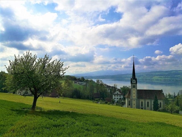 Blick auf eine grüne Wiese mit einer Kirche und einem See im Hintergrund. Der Himmel ist teilweise bewölkt.