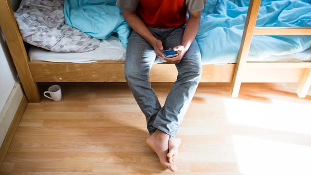 Mann in Jeans sitzt ohne Socken auf einem Bett, hat sein Handy in der Hand. Mann in Jeans sitzt ohne Socken auf einem Bett, hat sein Handy in der Hand.