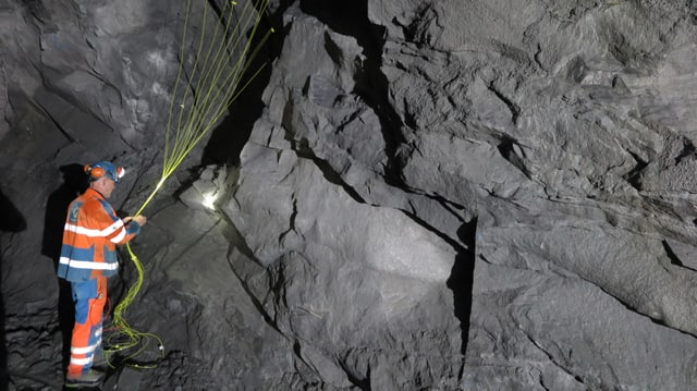 Ein Mann mir roter Leuchtweste und Stirnlampe steht in einem Tunnel im Felsen mit vielen gelben Zündschnüren in der Hand. Ein Mann mir roter Leuchtweste und Stirnlampe steht in einem Tunnel im Felsen mit vielen gelben Zündschnüren in der Hand.