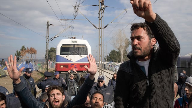 Flüchtlinge an der Grenze zu Mazedonien verlangen Durchlass, im Hintergrund eine Zugslokomotive Flüchtlinge an der Grenze zu Mazedonien verlangen Durchlass, im Hintergrund eine Zugslokomotive