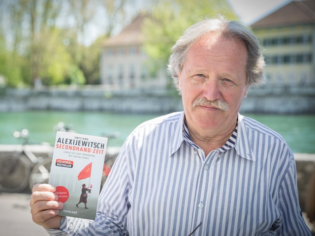 Ein Mann im Hemd steht am Ufer der Aare mit einem Buch in der Hand.