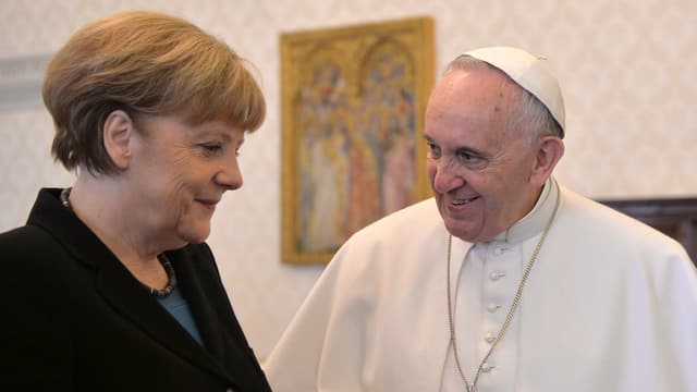 Merkel und der Papst stehen nebeneinander. Beide lächeln.