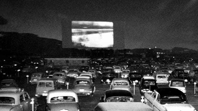 Ein altes schwarz-weiss-Foto: Auf einer Leinwand läuft ein Film, davor sind viele Autos prakiert. Ein altes schwarz-weiss-Foto: Auf einer Leinwand läuft ein Film, davor sind viele Autos prakiert.