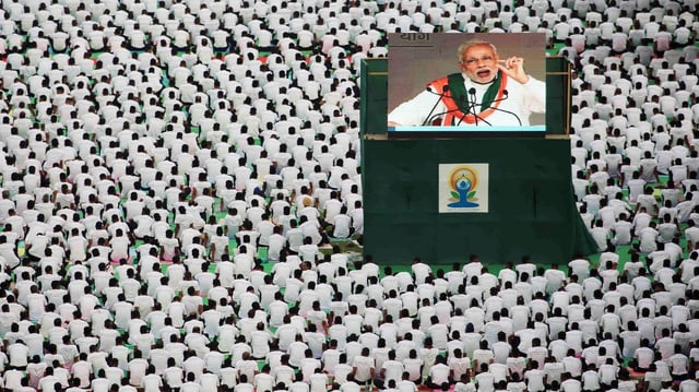 Wiese voll mit sitzenden, weiss gekleideten Menschen in Neu Dehli, in ihrer Mitte spricht auf einer Grossleinwand Premier Narendra Modi zu ihnen Wiese voll mit sitzenden, weiss gekleideten Menschen in Neu Dehli, in ihrer Mitte spricht auf einer Grossleinwand Premier Narendra Modi zu ihnen