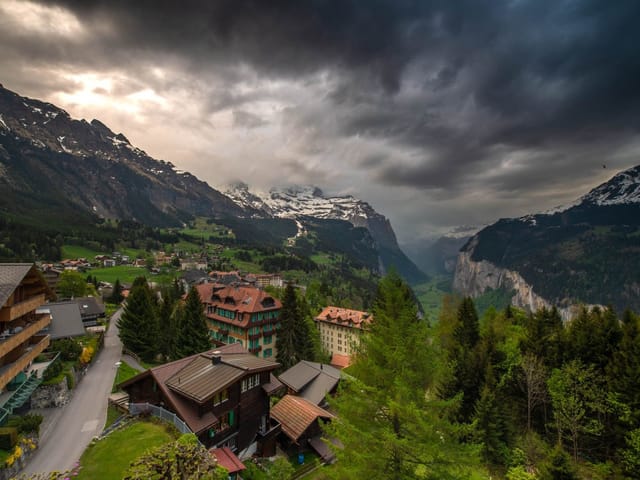 Bergwelt bei stark bewölktem Wetter mit einem Tal und einem Dorf am Hang. Die Wiesen sind schon grün.