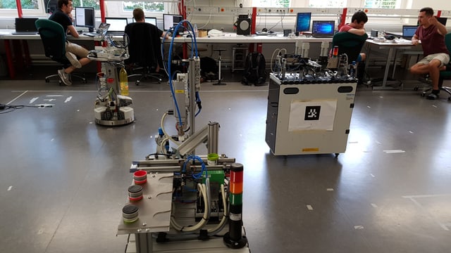 Ein Kasten und eine Maschine stehen auf einem Fussboden, im Hintergrund junge Männer am Computer. Ein Kasten und eine Maschine stehen auf einem Fussboden, im Hintergrund junge Männer am Computer.