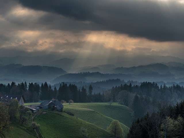 Hügelige Landschaft mit Wiesen und Wäldern, darüber spannt sich ein Himmel mit dunklen Wolken und hellen Lücken.