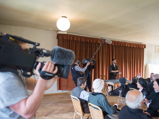 Gross im Vordergrund Kamera, hinten Leute am Tisch, Cedric Wermuth am Rednerpult