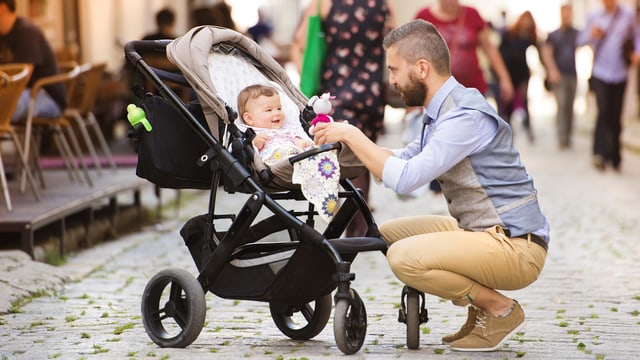 Ein Mann kniet auf der Strasse runter zu einem Kinderwagen und lächelt ein kleines Kind an. Ein Mann kniet auf der Strasse runter zu einem Kinderwagen und lächelt ein kleines Kind an.