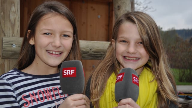 Annika und Sophie stehen im Freien und halten je ein Mikrofon in den Händen. Annika und Sophie stehen im Freien und halten je ein Mikrofon in den Händen.