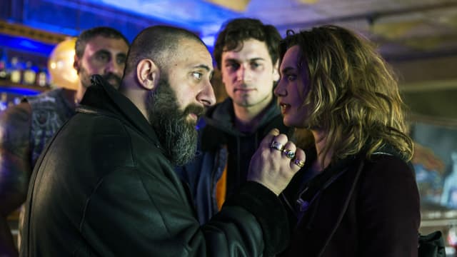 Ein Mann mit Bart und eine Frau stehen nahe voreinander und diskutieren. Im Hintergrund schauen zwei Männer zu, der eine hat ein verschlagenes Gesicht. Ein Mann mit Bart und eine Frau stehen nahe voreinander und diskutieren. Im Hintergrund schauen zwei Männer zu, der eine hat ein verschlagenes Gesicht.