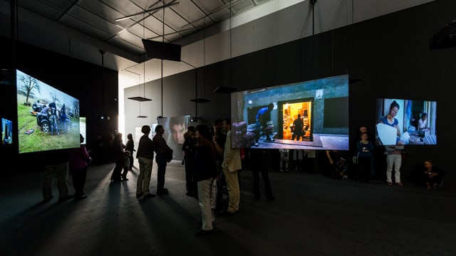 Menschen stehen vor Filmleinwänden in einem Ausstellungsraum. Menschen stehen vor Filmleinwänden in einem Ausstellungsraum.