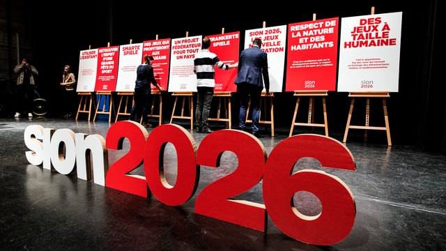 Plakate für Sion 2026 werden auf einer Bühne aufgestellt. Plakate für Sion 2026 werden auf einer Bühne aufgestellt.