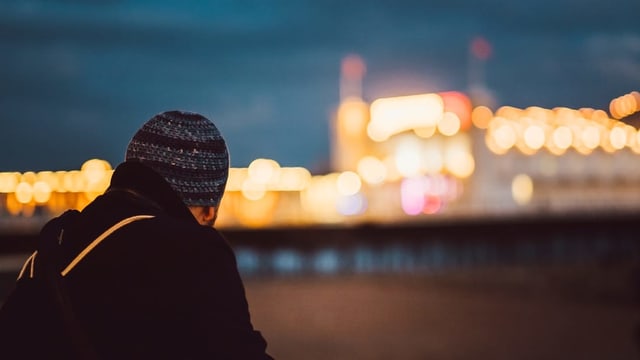 Ein junger Mann mit Kappe schaut aus der Ferne auf eine beleuchtete Stadt. Ein junger Mann mit Kappe schaut aus der Ferne auf eine beleuchtete Stadt.