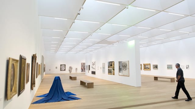 Ein Mann geht durch helle Ausstellungsräume mit Kunstwerken an den Wänden und am Boden Ein Mann geht durch helle Ausstellungsräume mit Kunstwerken an den Wänden und am Boden