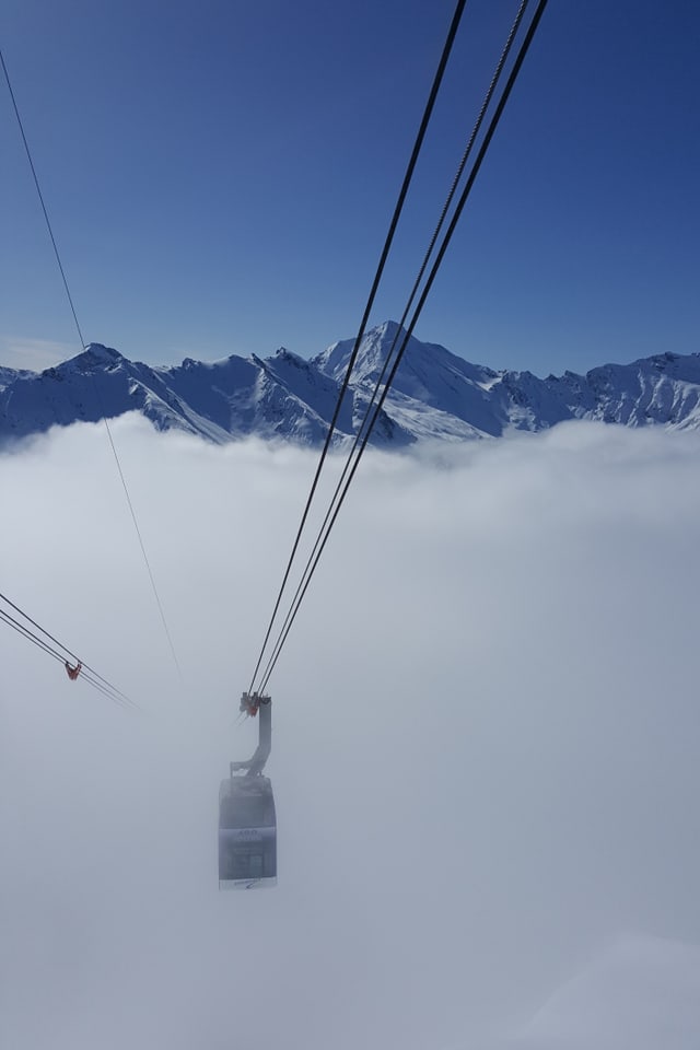Eine Seilbahn steigt aus dem Nebelgrau. Der Himmel über den Bergen erstrahlt tiefblau.