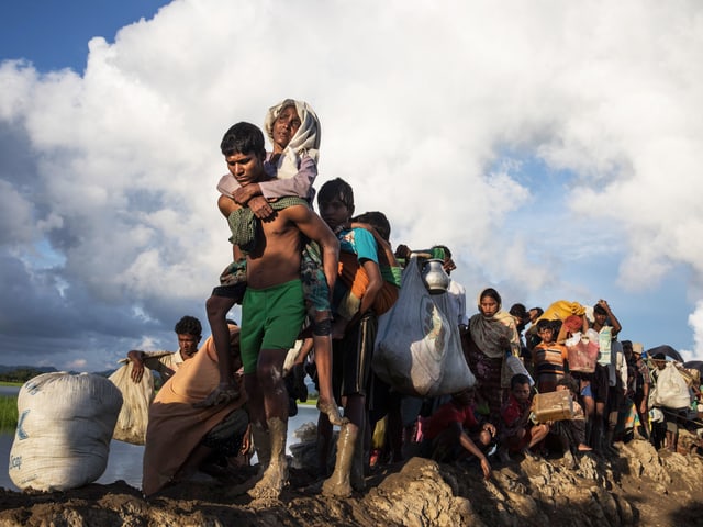 Rohingyas auf der Flucht: Eine Gruppe Menschen geht hintereinander, vorne trägt ein Mann eine alte Frau. Rohingyas auf der Flucht: Eine Gruppe Menschen geht hintereinander, vorne trägt ein Mann eine alte Frau.