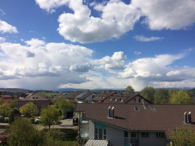 Mix aus Wolken und blauem Himmel. Im Vordergrund Häuser und Bäume.