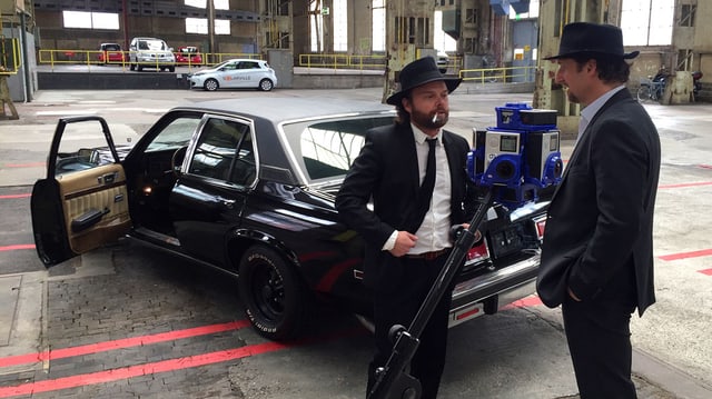 Auf dem Set: Zwei Männer stehen in einer Lagerhalle vor einem Auto und werden von einer 360-Grad-Kamera gefilmt. Auf dem Set: Zwei Männer stehen in einer Lagerhalle vor einem Auto und werden von einer 360-Grad-Kamera gefilmt.
