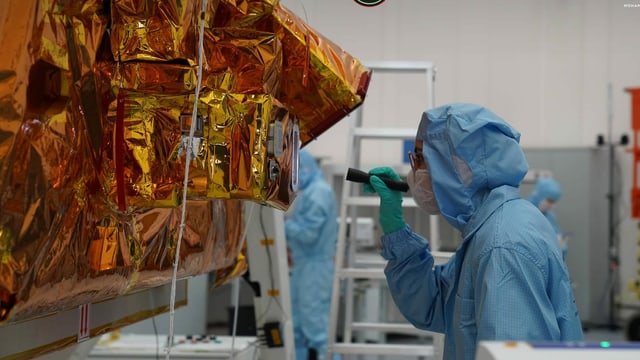 Eine Forscherin in Schutzkleidung arbeitet am Marssatelliten, der mit oranger Folie eingepackt ist. Eine Forscherin in Schutzkleidung arbeitet am Marssatelliten, der mit oranger Folie eingepackt ist.