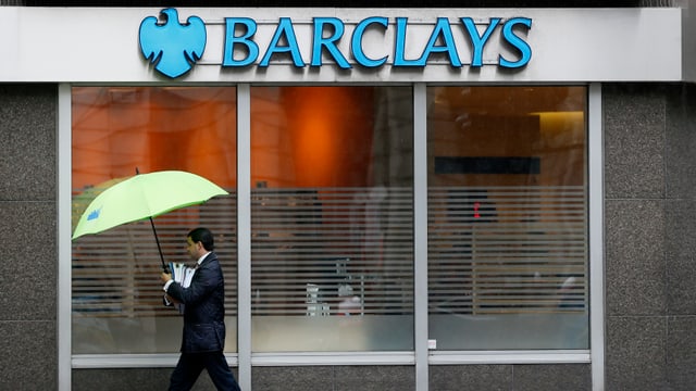 Ein Banker in Anzug und mit Regenschirm läuft an einer Filiale der Barclays Bank vorbei. Ein Banker in Anzug und mit Regenschirm läuft an einer Filiale der Barclays Bank vorbei.