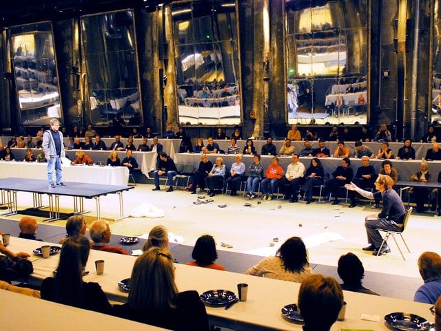 Menschen sitzen an langen Tischen, in der Mitte die Schauspieler, an den Wänden Spiegel Menschen sitzen an langen Tischen, in der Mitte die Schauspieler, an den Wänden Spiegel