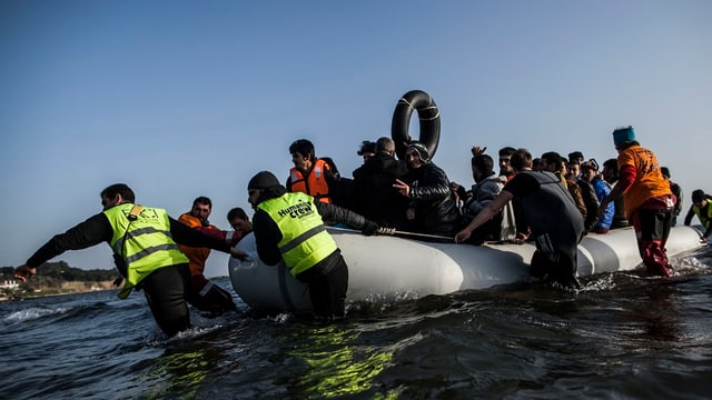 Mindestens acht Personen, eils mit gelben Signalwesten, bringen ein grosses Gummiboot, das voll ist mit Flüchtlingen, an Land. Mindestens acht Personen, eils mit gelben Signalwesten, bringen ein grosses Gummiboot, das voll ist mit Flüchtlingen, an Land.