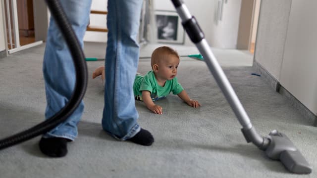Ein Kind am Boden, während eine erwachsene Person staubsaugt. Ein Kind am Boden, während eine erwachsene Person staubsaugt.