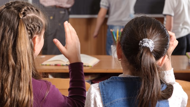 Zwei Mädchen sitzen auf der Schulbank und halten die Handhoch. Zwei Mädchen sitzen auf der Schulbank und halten die Handhoch.