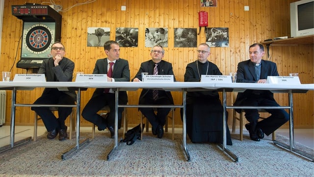 Fünf Männer sitzen an einem langen Tisch. Im Hintergrund hängen Fotos an der Wand. Fünf Männer sitzen an einem langen Tisch. Im Hintergrund hängen Fotos an der Wand.
