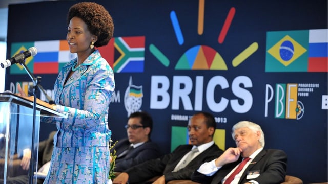 Frau an einem Rednerpult, dahinter Männer auf Stühlen und Brics-Logo. Frau an einem Rednerpult, dahinter Männer auf Stühlen und Brics-Logo.