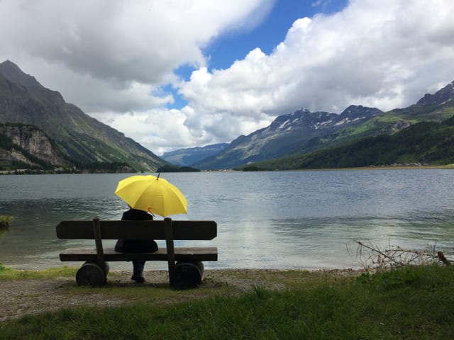 Bergsee mit Bank. Eine Person mit gelbem Regenschirm sitzt darauf. Sie blickt auf die Berge und den teils bewölkten Himmel.