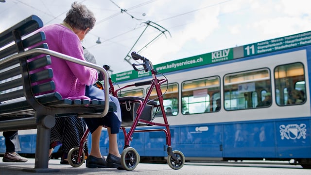 Frau auf einer Bank mit Rollator, vor ihr fährt ein blaues Tram vorbei. Frau auf einer Bank mit Rollator, vor ihr fährt ein blaues Tram vorbei.