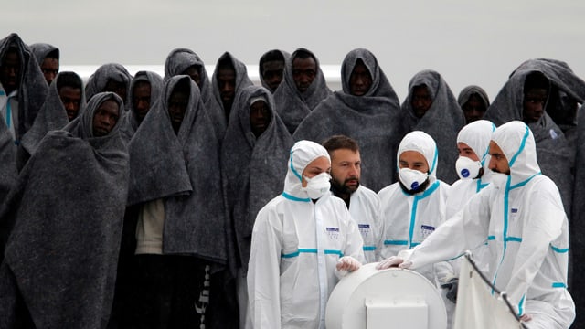 Flüchtlinge in dunkelgraue Wolldecken gehüllt stehen im Hintergrund. Im Vordergrund Helfer in weissen Schutzanzügen mit Mundschutz. Flüchtlinge in dunkelgraue Wolldecken gehüllt stehen im Hintergrund. Im Vordergrund Helfer in weissen Schutzanzügen mit Mundschutz.