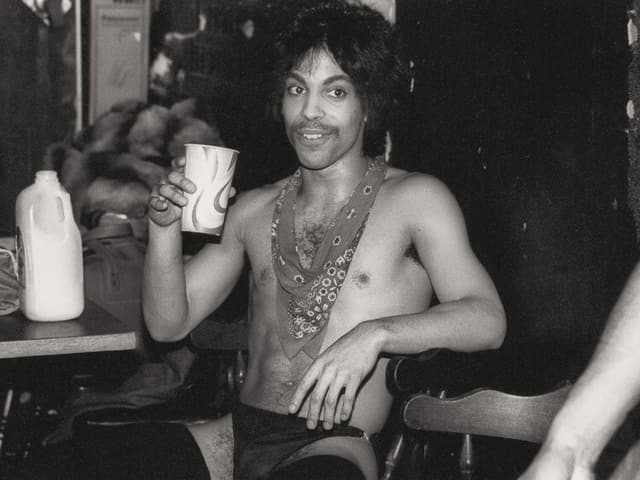 Ein Foto von Prince von 1985: Er sitzt halbnackt in einem Stuhl und trinkt aus einem Becher. Ein Foto von Prince von 1985: Er sitzt halbnackt in einem Stuhl und trinkt aus einem Becher.