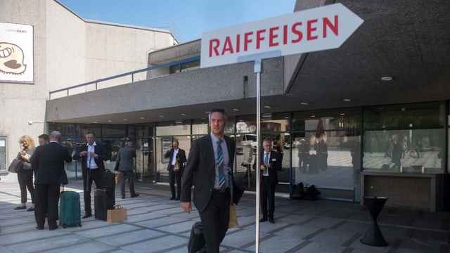 Raiffeisen-Wegweiser, dahinter Leute in Anzügen vor einem Bau. Raiffeisen-Wegweiser, dahinter Leute in Anzügen vor einem Bau.