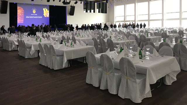 Ein Saal mit weiss gedeckten Tischen, im Hintergrund Menschen vor einer Leinwand Ein Saal mit weiss gedeckten Tischen, im Hintergrund Menschen vor einer Leinwand
