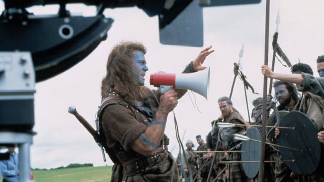 Ein Mann mit langen Haaren, Schottenrock und einem Megafon in der Hand steht vor einer Horde verkleideter Männer und gibt Anweisungen. Ein Mann mit langen Haaren, Schottenrock und einem Megafon in der Hand steht vor einer Horde verkleideter Männer und gibt Anweisungen.