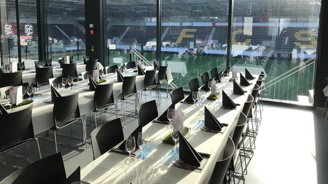 Stadion-Restaurant mit weissen Tischen und schwarzen Stühlen, dahinter Blick auf Tribüne und Rasen Stadion-Restaurant mit weissen Tischen und schwarzen Stühlen, dahinter Blick auf Tribüne und Rasen